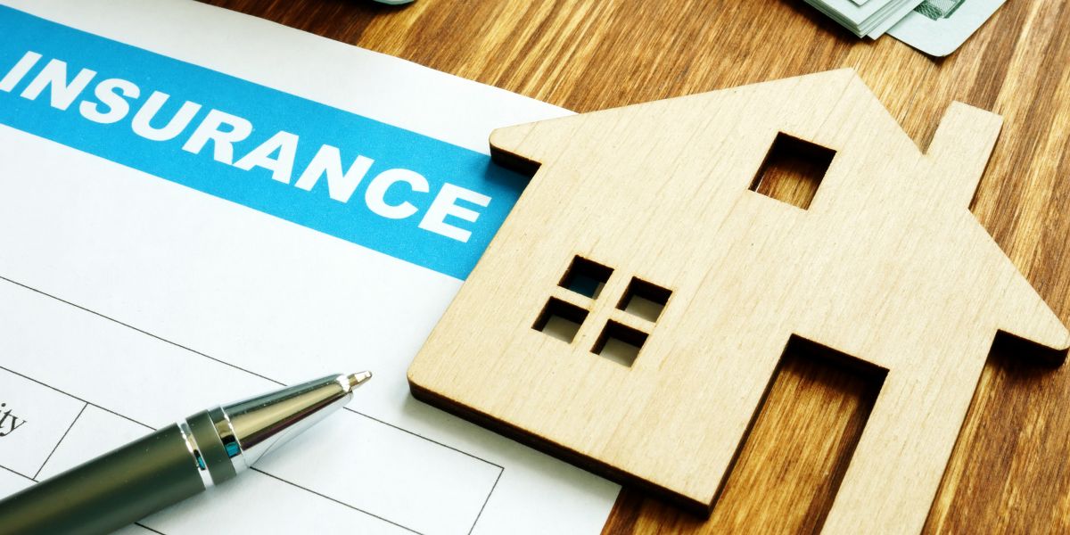 Wooden house model on top of a home insurance document with a pen nearby.