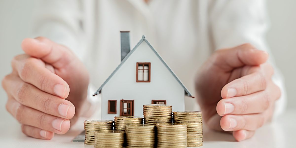 Hands protecting a small house model surrounded by stacks of coins.